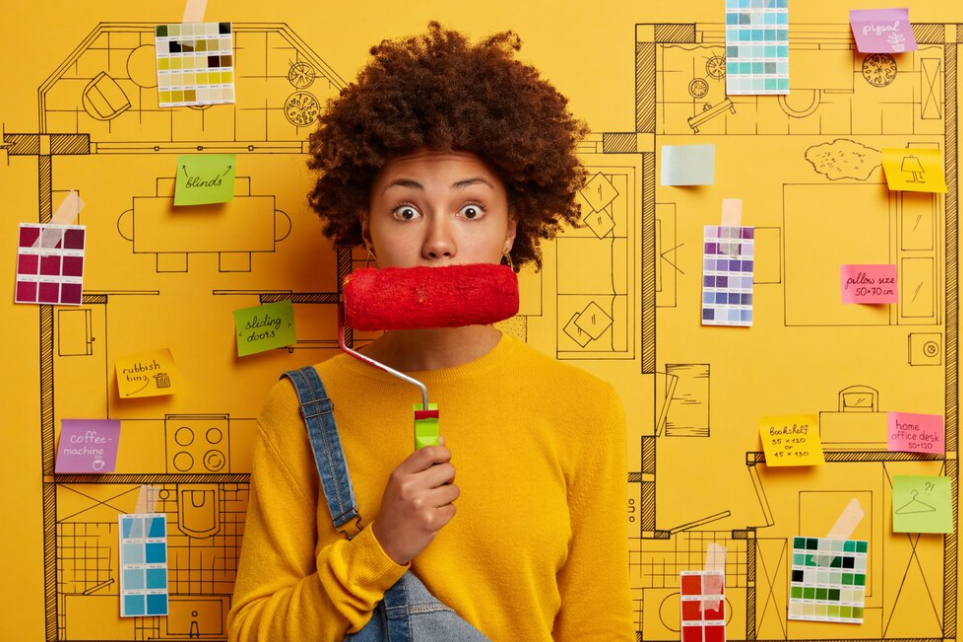 woman in a yellow sweater holds a platen near the mouse, a yellow wall behind with notes and color pallets pinned on it