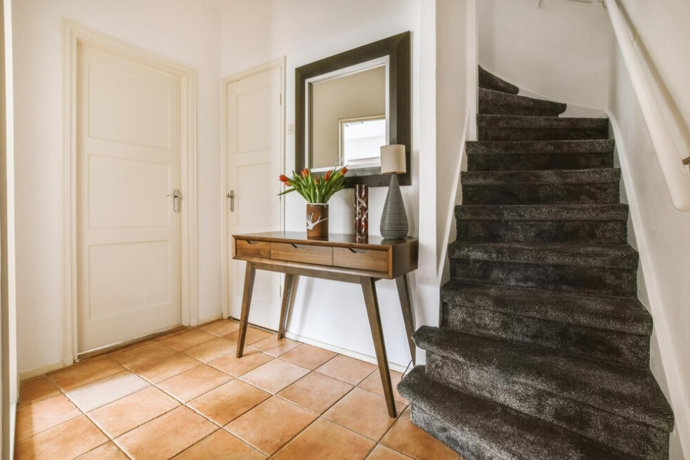 corridor with tiled floors, a table with a vase and a mirror above it, and a black carpeted staircase