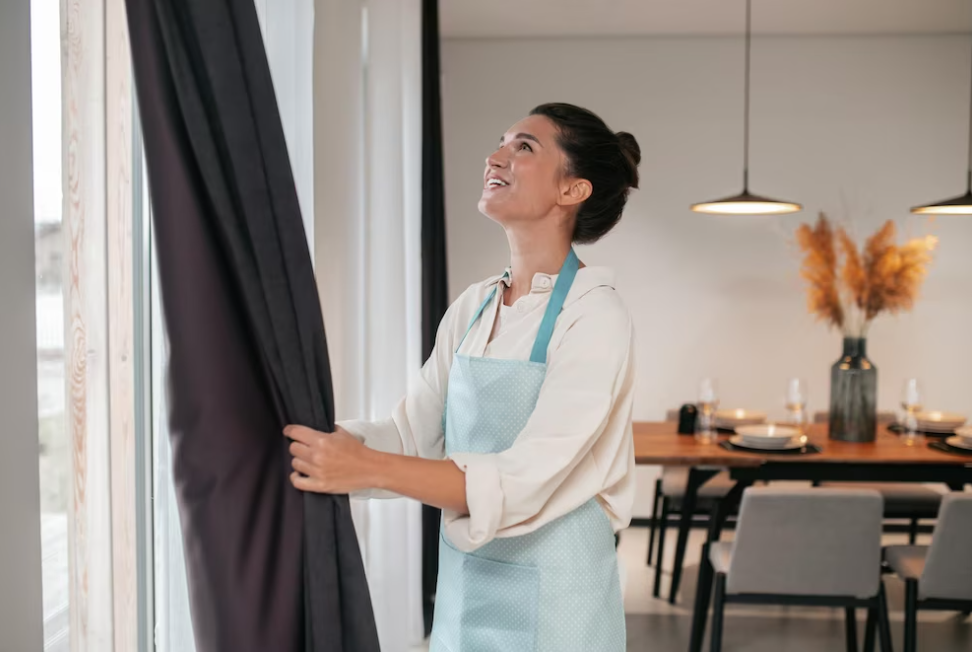 woman standing near the window and holding the curtains in her hands, table with vase and gray chairs near it